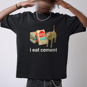 May include: Black t-shirt with a graphic of a cat standing next to a stack of cement bags. The text "i eat cement" is printed below the image.