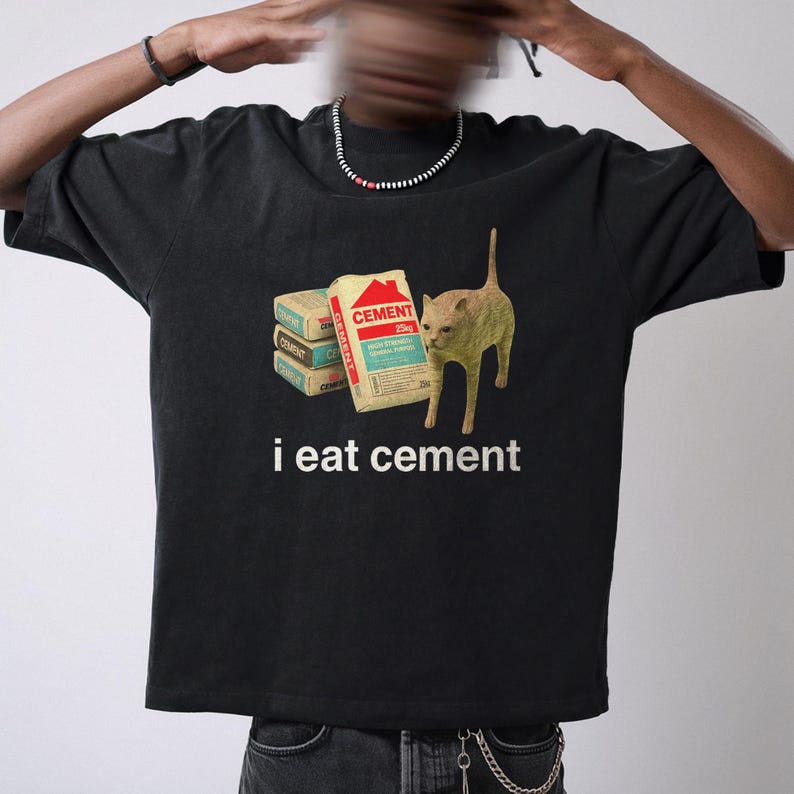 May include: Black t-shirt with a graphic of a cat standing next to a stack of cement bags. The text "i eat cement" is printed below the image.