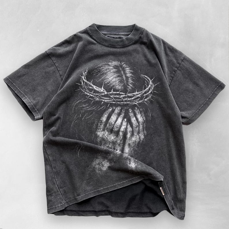 May include: Dark gray t-shirt featuring a distressed graphic of a person with a crown of thorns and hands covering their face. The design is in white, creating a striking contrast against the shirt's dark background. The shirt has short sleeves and a crew neck.