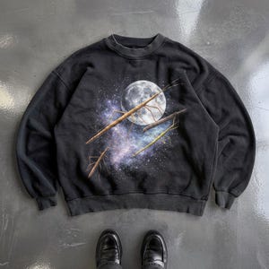 May include: A black crewneck sweatshirt featuring a graphic print of three stick insects on branches against a backdrop of a full moon and a purple and blue galaxy nebula. The long-sleeved garment has a ribbed collar, cuffs, and hem.