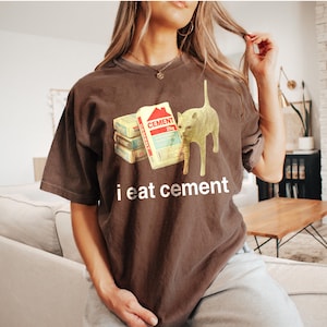 May include: A brown t-shirt with a graphic of a cat eating a stack of cement bags. The text "i eat cement" is printed below the graphic.