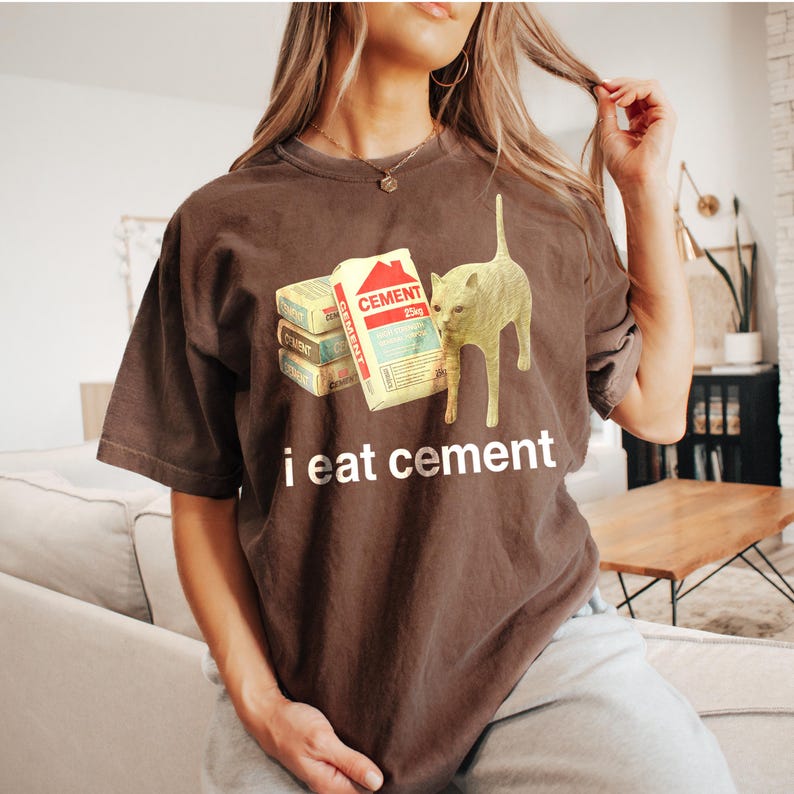 May include: A brown t-shirt with a graphic of a cat eating a stack of cement bags. The text "i eat cement" is printed below the graphic.