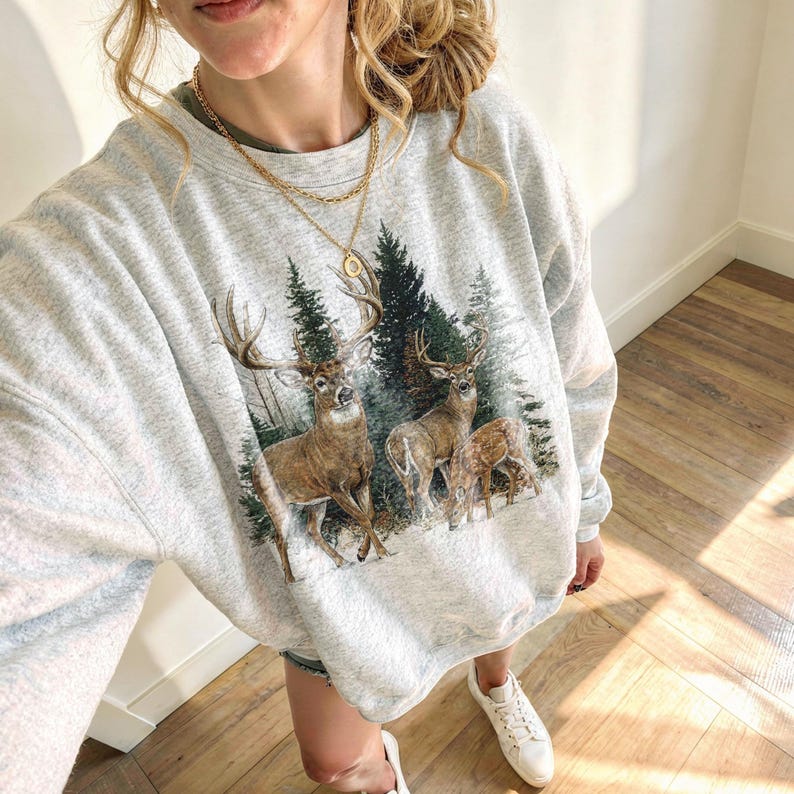 May include: A light grey crew neck sweatshirt with a forest and deer design. The print depicts a buck with antlers, two other deer, and fir trees. The sweatshirt is a casual style.