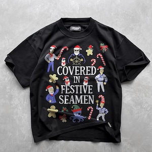 May include: Black crew neck t-shirt with a festive nautical design. The graphic includes cartoon sailors, penguins, candy canes, and the text "COVERED IN FESTIVE SEAMEN". The design incorporates red, white, blue, and yellow colours.