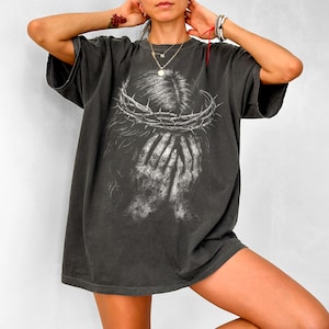 May include: Dark gray oversized t-shirt featuring a graphic of a crown of thorns and hands covering a face. The design is in white, creating a striking contrast against the shirt's color. The shirt has short sleeves and a relaxed fit.