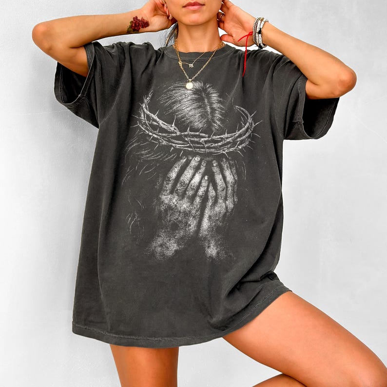 May include: Dark gray oversized t-shirt featuring a graphic of a crown of thorns and hands covering a face. The design is in white, creating a striking contrast against the shirt's color. The shirt has short sleeves and a relaxed fit.