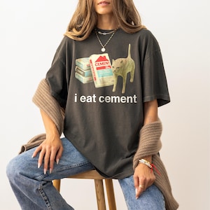 May include: A dark gray t-shirt with a graphic of a cat looking at a stack of cement bags. The text "i eat cement" is printed below the graphic.