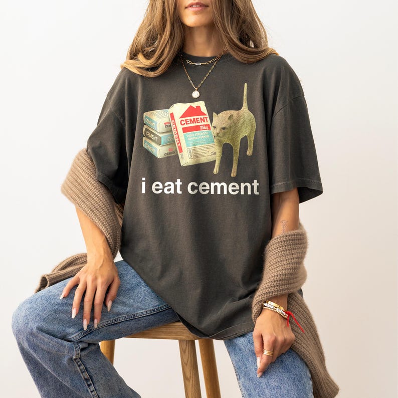 May include: A dark gray t-shirt with a graphic of a cat looking at a stack of cement bags. The text "i eat cement" is printed below the graphic.