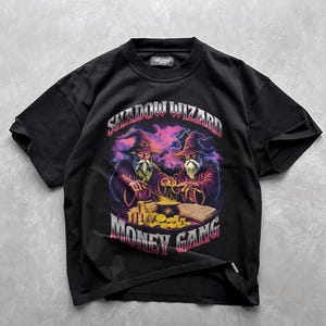 May include: Black t-shirt with a graphic print of two wizards with purple robes and pointed hats. The wizards are holding a cauldron and a book. The text "SHADOW WIZARD MONEY GANG" is printed in white and pink on the shirt.