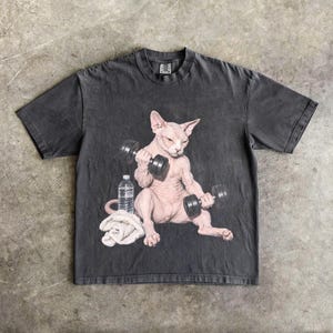 May include: A charcoal grey short-sleeve t-shirt featuring a graphic print of a hairless cat lifting two black dumbbells. Next to the cat is a clear plastic water bottle and a folded white towel. The shirt has a crew neckline and a vintage wash.