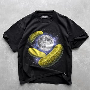 Puede incluir: Una camiseta negra de manga corta con un estampado gráfico de tres pepinillos verdes flotando en el espacio alrededor de una luna blanca detallada sobre un fondo de nebulosa estrellada en tonos morados y azules. Tiene cuello redondo.