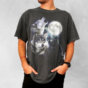 K&ouml;nnte beinhalten: Dunkelgraues T-Shirt mit einer Grafik von drei W&ouml;lfen, die einen Vollmond anheulen. Die W&ouml;lfe sind in Grau- und Schwarzt&ouml;nen dargestellt, mit dem Mond im Hintergrund. Das T-Shirt hat einen Vintage-Look.