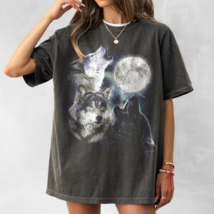 K&ouml;nnte beinhalten: Dunkelgraues T-Shirt mit einer Grafik von drei W&ouml;lfen, die den Vollmond anheulen. Das Design umfasst detaillierte Illustrationen der W&ouml;lfe und einen hellen Mond vor einem Sternenhintergrund. Das Shirt hat einen Vintage-Look.