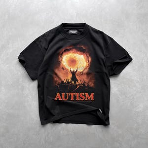 Puede incluir: Camiseta negra con un estampado gr&aacute;fico de una persona de pie frente a una explosi&oacute;n de fuego. El texto "AUTISM" est&aacute; impreso en rojo debajo del gr&aacute;fico.