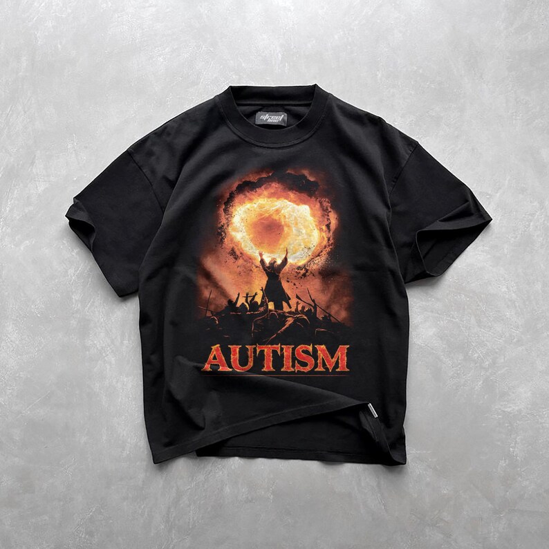 Puede incluir: Camiseta negra con un estampado gr&aacute;fico de una persona de pie frente a una explosi&oacute;n de fuego. El texto "AUTISM" est&aacute; impreso en rojo debajo del gr&aacute;fico.