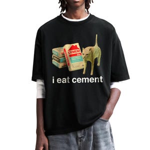 May include: Black t-shirt with a white cat and bags of cement printed on the front. The text "i eat cement" is printed below the image.