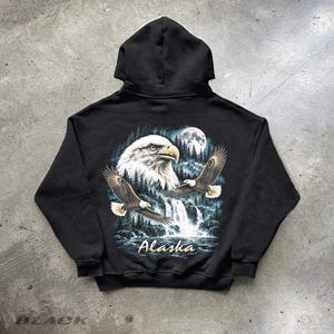 Op de afbeelding: Een zwarte hoodie met een grafische print op de achterkant. Het ontwerp toont een grote kop van een Amerikaanse zeearend, een vliegende adelaar, een waterval, een bos en een volle maan. Het woord Alaska staat in een lichtgekleurd cursief lettertype.