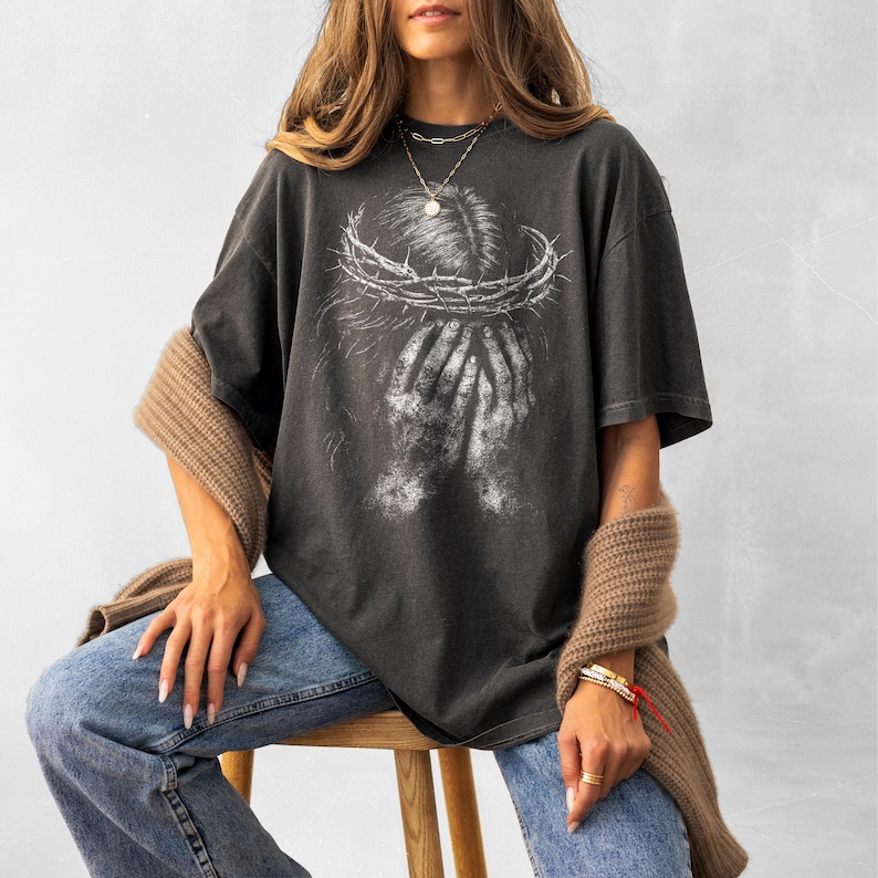 May include: Dark gray t-shirt featuring a distressed graphic of a crown of thorns above hands covering a face. The shirt has a vintage, worn look. The model is wearing blue jeans and a brown sweater.