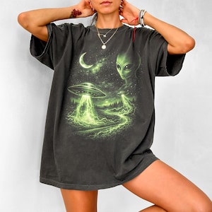 Könnte beinhalten: Ein anthrazitfarbenes Kurzarm-T-Shirt mit einem Grafikdruck eines grünen Alien-Kopfes, einer Mondsichel und zwei fliegenden Untertassen, die hellgrüne Lichtstrahlen über eine dunkle Waldlandschaft mit einem sternenklaren Nachthimmel aussenden.