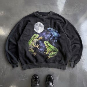 Può includere: Una felpa nera a girocollo con una stampa grafica raffigurante due rane verdi e una rana blu su uno sfondo con una luna piena luminosa e un cielo stellato viola. Il capo presenta maniche lunghe e colletto e polsini a coste.