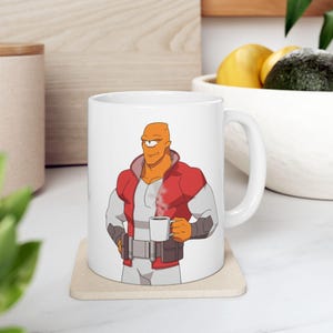 May include: White ceramic mug featuring a cartoon character with orange skin, one eye, and a red and white jacket. The character is holding a steaming cup of coffee. The mug is on a beige coaster.