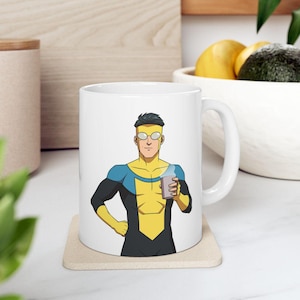 May include: White ceramic mug featuring a cartoon illustration of a superhero character in a yellow and blue costume, holding a cup. The mug is placed on a beige coaster.