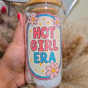 May include: A clear glass tumbler with a bamboo lid and straw. The tumbler features a colourful design with the text "HOT GIRL ERA" in pink and blue, surrounded by flowers and stars.
