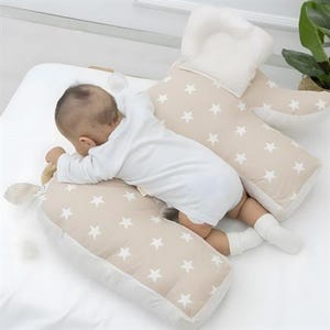 May include: A baby rests on a beige pillow set with white star patterns. The set includes a headrest and two body pillows. The baby wears a white onesie and socks. The pillows have a striped border. A wooden toy is in the baby's hand.