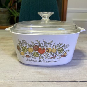 May include: White casserole dish with a glass lid. The dish has a floral design with vegetables and the text "L'Echalote La Marjolaine".