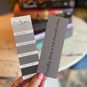 May include: A gray paint color chart with the text "MorallyGREY" at the top. The chart shows five different shades of gray with corresponding names and numbers. A separate gray card with the text "Morally grey is my favorite color" is held in a hand.