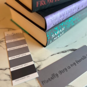 May include: A stack of three books with the titles "From Blood and Ash", "The Ballad of Never After", and "Sarah J. Maas" visible. A gray card with the text "Morally grey is my favorite" is in the foreground.