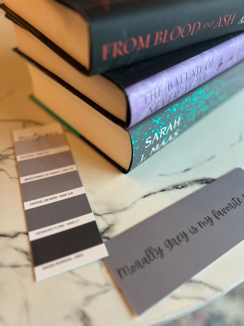 May include: A stack of three books with the titles "From Blood and Ash", "The Ballad of Never After", and "Sarah J. Maas" visible. A gray card with the text "Morally grey is my favorite" is in the foreground.