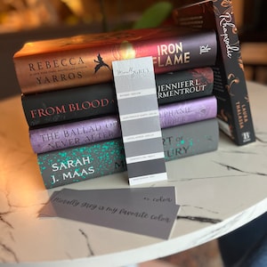 May include: A stack of fantasy novels with colorful covers, including "Iron Flame" by Jennifer L. Armentrout, "From Blood and Ash" by Jennifer L. Armentrout, "The Ballad of Never After" by Stephanie Garber, and "A Court of Thorns and Roses" by Sarah J. Maas. A paint chip labeled "Morally Grey" is on top of the books.
