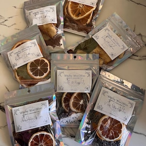 May include: Nine small, clear plastic bags with silver foil accents, each containing dried fruit, herbs, and spices. Each bag has a white label with the text "Witchy Woo Woo" and a different intention, such as "Abundance & Love", "Success & Happiness", "New Opportunities", "Positive Energy", "Wealth & Prosperity", and "Divine Protection".