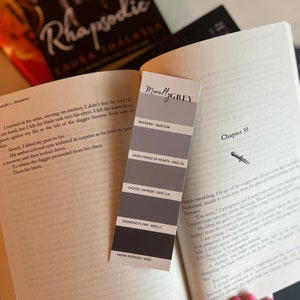 May include: A book titled "Rhapsodic" by Laura Thalassa with a bookmark featuring a color palette with the title "Morally Grey" and the names of different colors with their corresponding codes. The bookmark is placed on the page with the chapter title "Chapter 39".