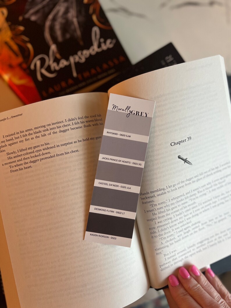 May include: A book titled "Rhapsodic" by Laura Thalassa with a bookmark featuring a color palette with the title "Morally Grey" and the names of different colors with their corresponding codes. The bookmark is placed on the page with the chapter title "Chapter 39".