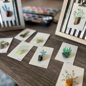 Puede incluir: Una colección de pequeñas piedras pintadas con varias plantas y flores. Las piedras se muestran en tarjetas blancas con las palabras "Art 21" escritas en ellas.