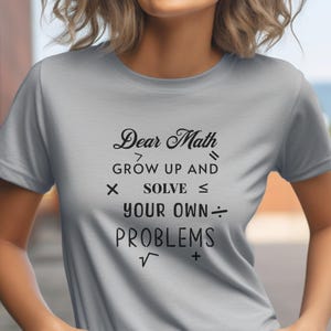 Könnte beinhalten: Ein graues T-Shirt mit einem schwarzen Aufdruck, der "Liebe Mathe, werde erwachsen und löse deine eigenen Probleme" lautet. Der Aufdruck enthält mathematische Symbole wie größer als, kleiner als, multiplizieren, dividieren und Quadratwurzel.