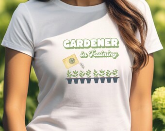 Può includere: Maglietta bianca con il testo "GARDENER in TRAINING" in lettere verdi. Un'immagine di un pacchetto di semi che versa semi in un vassoio di piantine.