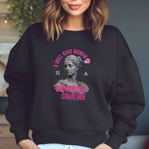 Peut inclure: Un sweat-shirt noir avec un imprimé graphique rose qui dit "Une femme bien lue est une créature dangereuse". Le graphique représente un buste de femme avec une pile de livres au-dessus de sa tête.