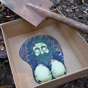May include: A mouse pad featuring a stylised illustration of a bearded, green-skinned forest figure with leafy hair and branches, integrated with two rounded, pale yellow wrist rests, all contained within a plain, open cardboard box.