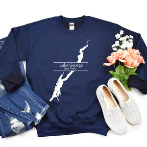 May include: Navy blue sweatshirt featuring a white outline of Lake George, New York. The sweatshirt is laid flat on a white background with a pair of blue jeans and white shoes.