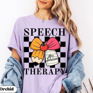 May include: Orchid purple t-shirt with a graphic design for speech therapy. The design features a pencil, bow, apple, and the text "SPEECH THERAPY" in black. A notepad with the name "Mrs. Johnson" is also included.