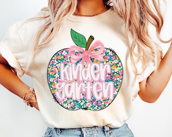 Puede incluir: Camiseta color crema con un diseño de manzana grande. La manzana está llena de un estampado floral y tiene un lazo rosa, una hoja verde y un tallo marrón. La palabra "Kindergarten" está en rosa. La camiseta es ropa casual.