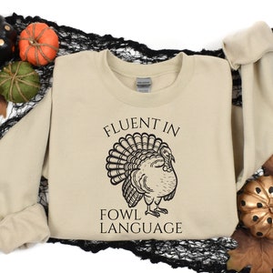May include: A beige sweatshirt with the text "FLUENT IN FOWL LANGUAGE" and a black illustration of a turkey. The sweatshirt is surrounded by decorative pumpkins and fall foliage.