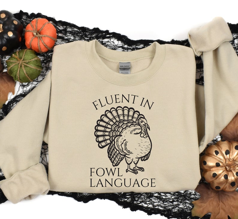 May include: A beige sweatshirt with the text "FLUENT IN FOWL LANGUAGE" and a black illustration of a turkey. The sweatshirt is surrounded by decorative pumpkins and fall foliage.