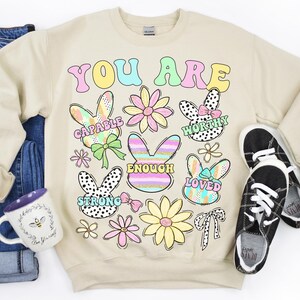 May include: Beige sweatshirt with the text "YOU ARE" in colorful letters. The sweatshirt features a variety of colorful bunny illustrations with words like "CAPABLE", "WORTHY", "ENOUGH", "LOVED", and "STRONG" inside each bunny.