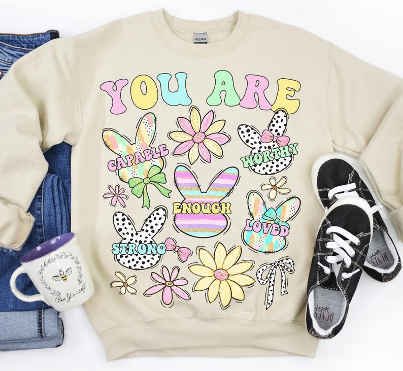 May include: Beige sweatshirt with the text "YOU ARE" in colorful letters. The sweatshirt features a variety of colorful bunny illustrations with words like "CAPABLE", "WORTHY", "ENOUGH", "LOVED", and "STRONG" inside each bunny.