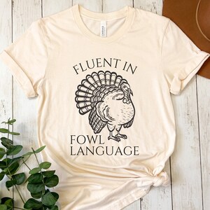 May include: A cream-coloured t-shirt featuring the text "FLUENT IN FOWL LANGUAGE" above a black illustration of a turkey. The shirt is made of a soft material and has rolled-up sleeves.