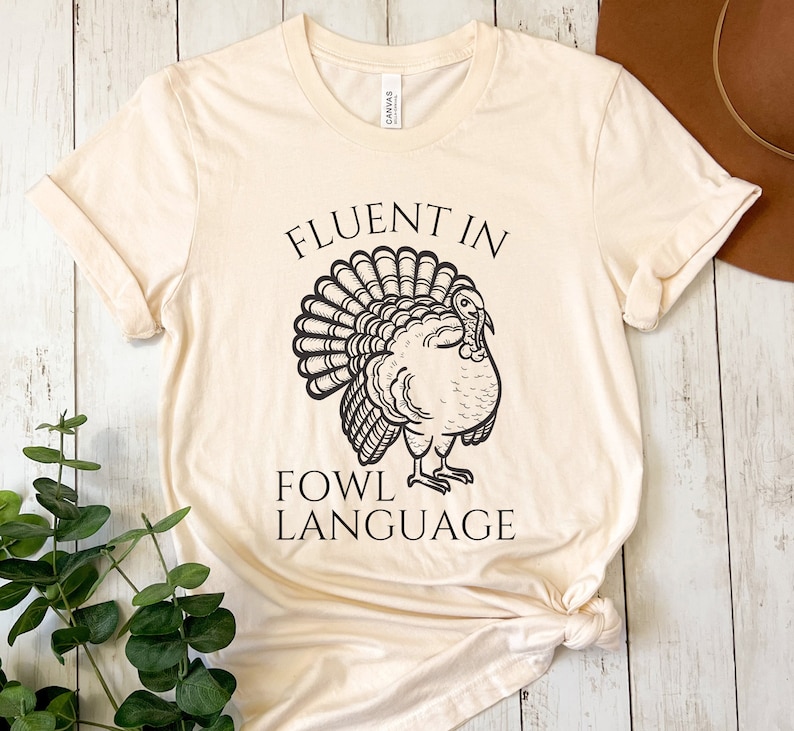 May include: A cream-coloured t-shirt featuring the text "FLUENT IN FOWL LANGUAGE" above a black illustration of a turkey. The shirt is made of a soft material and has rolled-up sleeves.
